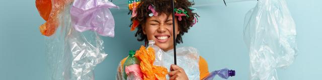 Emotional dark skinned woman stands under plastic umbrella, smirks face and shows white teeth, demonstrates environment awareness, wrapped in polyethylene film, isolated on blue. Zero waste concept