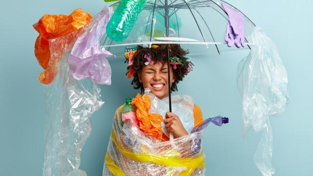 Emotional dark skinned woman stands under plastic umbrella, smirks face and shows white teeth, demonstrates environment awareness, wrapped in polyethylene film, isolated on blue. Zero waste concept