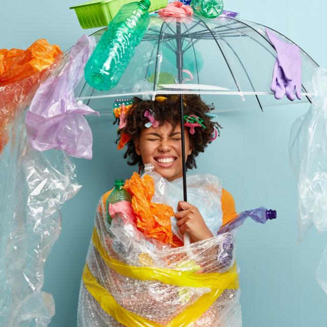 Emotional dark skinned woman stands under plastic umbrella, smirks face and shows white teeth, demonstrates environment awareness, wrapped in polyethylene film, isolated on blue. Zero waste concept